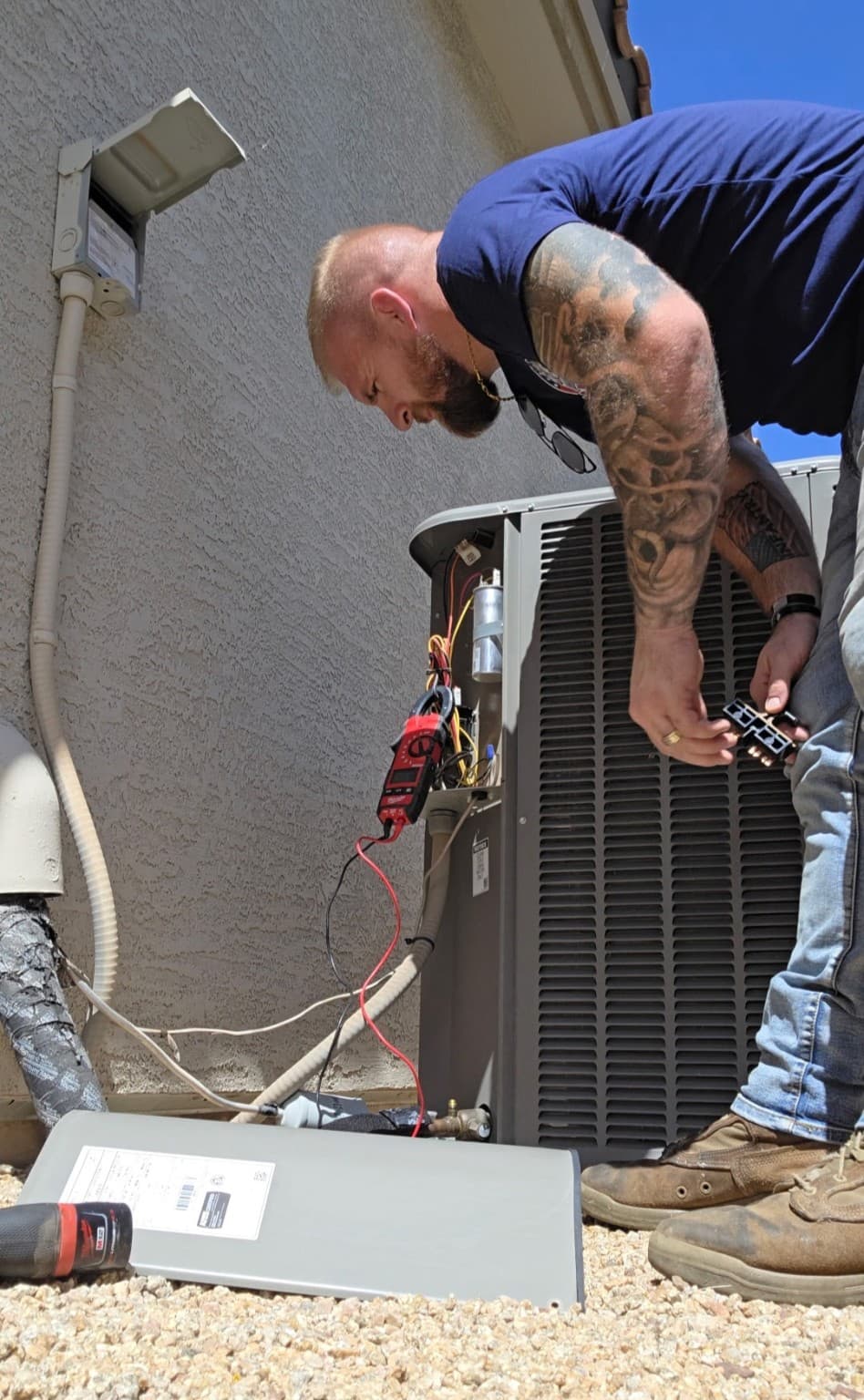 Tattooed technician uses a multimeter to inspect an outdoor air conditioning unit's electrical components.