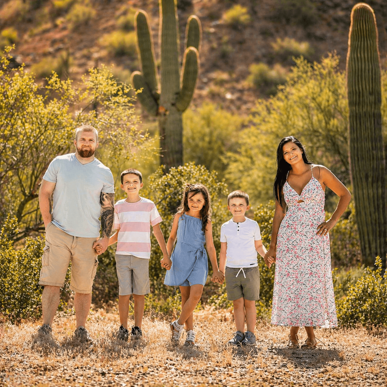 Smiling family of five holding hands in a sunny desert setting with tall saguaro cacti.